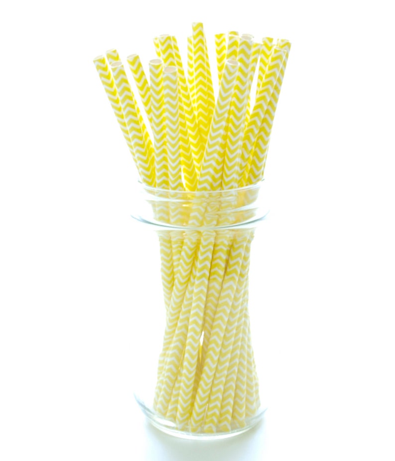May include: A collection of yellow and white chevron patterned paper straws in a clear glass. The straws are arranged vertically, with some extending above the rim of the glass. The background is plain white.