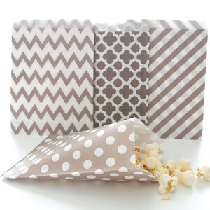 May include: A collection of decorative paper treat bags in various patterns: chevron, quatrefoil, and diagonal stripes. One bag is open, spilling popcorn. The bags are taupe and white, suitable for party favors or snacks.