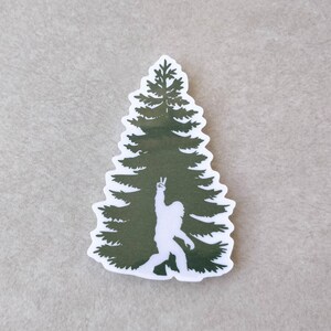 Puede incluir: Una silueta verde de un pino con una silueta blanca de un Bigfoot dentro del &aacute;rbol, haciendo un signo de paz con su mano.