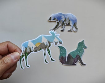 Könnte beinhalten: Drei weiße Silhouetten-Aufkleber eines Bären, Elchs und Fuchses mit einem Aquarell-Landschaftsdesign in jeder Silhouette. Der Bär blickt nach links, der Elch nach rechts und der Fuchs nach rechts.