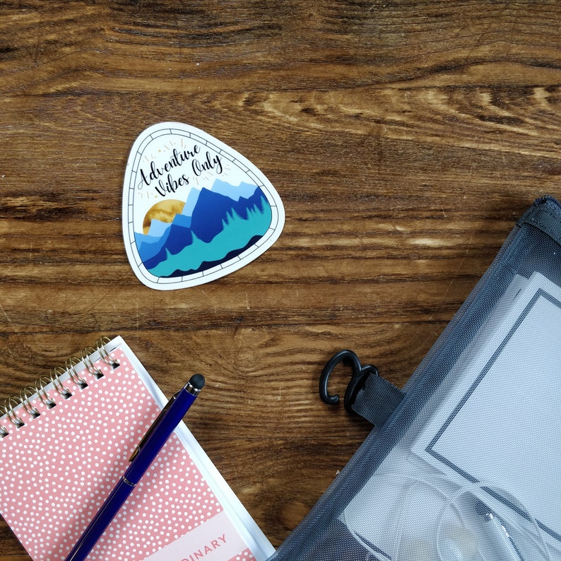 Op de afbeelding: Een sticker met een bergketen en een zon, met de tekst "Adventure Vibes Only". De sticker zit op een houten oppervlak naast een roze notitieboekje met witte stippen en een blauwe pen.