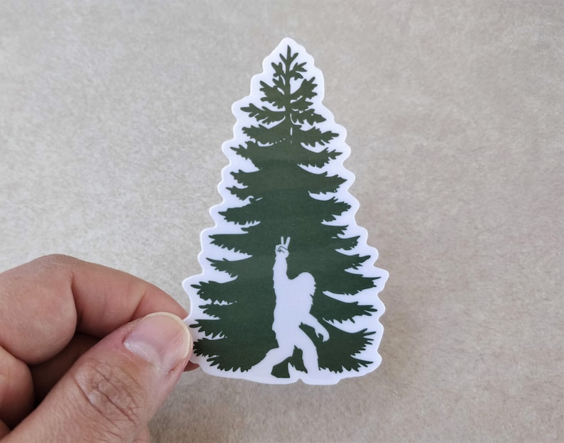 Puede incluir: Una silueta verde de un pino con una silueta blanca de Bigfoot dentro del &aacute;rbol, haciendo un signo de paz con su mano.