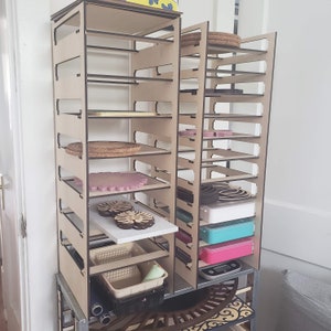 May include: A wooden storage rack with multiple shelves, holding various items. The shelves display round cork coasters, pink and brown decorative pieces, and containers. The rack is made of light-colored wood with dark edges.