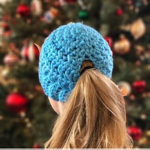 May include: A blue crochet hat with a ponytail hole in the back. The hat is worn by a person with long blonde hair. The text "CROCHET PATTERN from Made with a Twist" is displayed below the hat.
