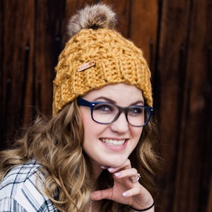 May include: A woman wearing a yellow crochet beanie with a brown pom pom and glasses. The beanie has a brown leather tag with the text "Made with a Twist" on it. The text "CROCHET PATTERN from Made with a Twist" is on a white banner below the image.