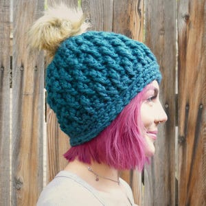 Puede incluir: Gorro tejido de color azul verdoso con un patrón texturizado y un pompón marrón claro y esponjoso en la parte superior. El gorro está hecho de hilo grueso y parece cálido y acogedor. Un elegante accesorio de invierno.