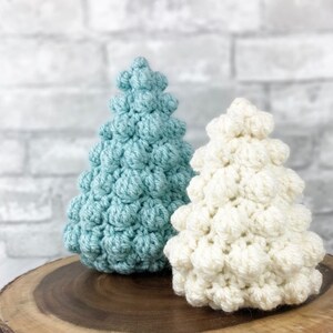 May include: Two crocheted Christmas trees, one in a light blue color and the other in white, are displayed on a wooden surface.