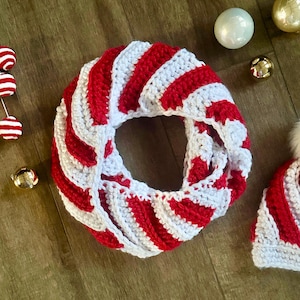 Peut inclure: &Eacute;charpe infinie au crochet avec un motif ray&eacute; rouge et blanc. L'&eacute;charpe est circulaire et semble douce et chaude. Des d&eacute;corations de No&euml;l sont &eacute;galement visibles, sugg&eacute;rant un th&egrave;me de saison. Un bonnet assorti est &eacute;galement visible.