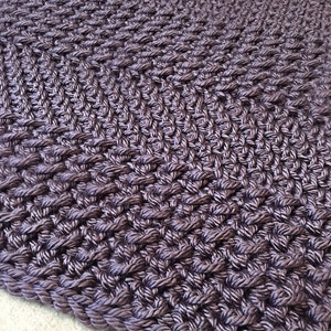 May include: A close-up of a dark purple braided rug with a diamond pattern. The rug is made of thick, textured rope and has a soft, plush feel.