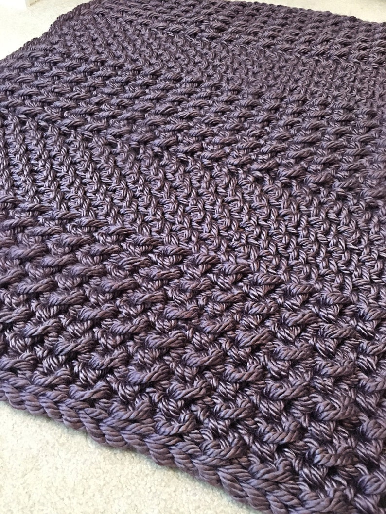 May include: A close-up of a dark purple braided rug with a diamond pattern. The rug is made of thick, textured rope and has a soft, plush feel.