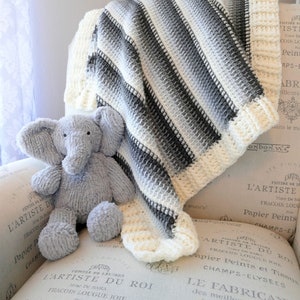 May include: A white and gray striped crocheted baby blanket with a gray crocheted elephant plush toy on top of it. The blanket is folded and sitting on a white and gray patterned surface.