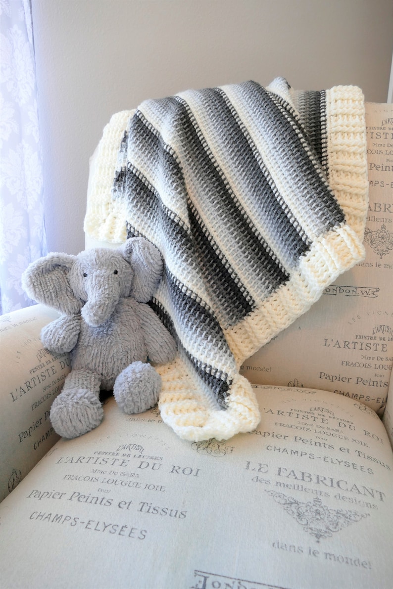 May include: A white and gray striped crocheted baby blanket with a gray crocheted elephant plush toy on top of it. The blanket is folded and sitting on a white and gray patterned surface.