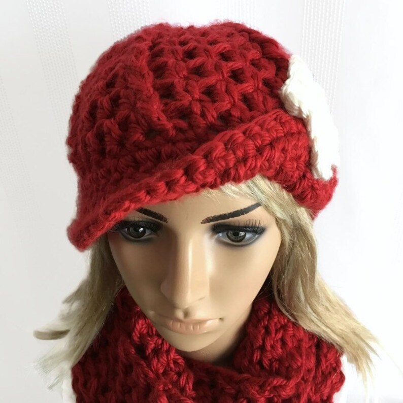 May include: A red crocheted hat with a small brim and a white accent. The hat is paired with a matching red scarf. The handmade hat and scarf set is a cozy accessory.