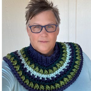 May include: A woman wearing a blue and green crocheted cowl with a navy blue turtleneck. The cowl has a geometric pattern with stripes of green, blue, and white.