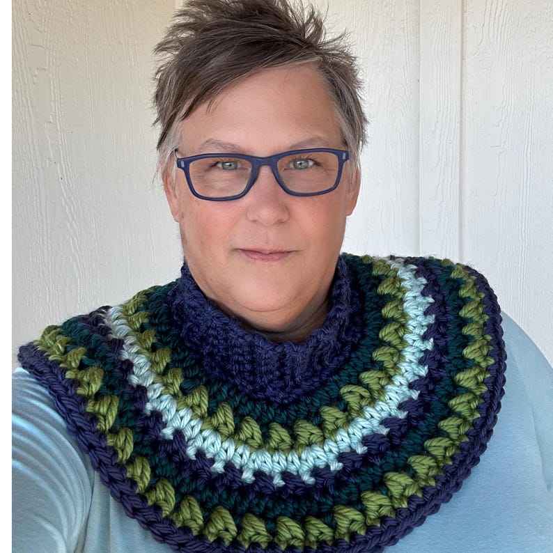 May include: A woman wearing a blue and green crocheted cowl with a navy blue turtleneck. The cowl has a geometric pattern with stripes of green, blue, and white.