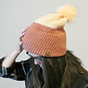 May include: A pink and cream crocheted beanie with a fluffy pom pom on top. The beanie has a brown leather tag with the word "Cozy" on it.