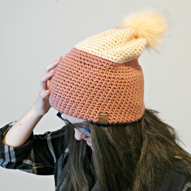 May include: A pink and cream crocheted beanie with a fluffy pom pom on top. The beanie has a brown leather tag with the word "Cozy" on it.