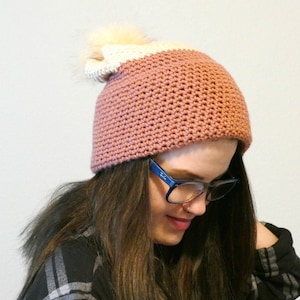 May include: A pink and white crocheted beanie with a fluffy white pom pom. The beanie is worn by a person with long brown hair and glasses.