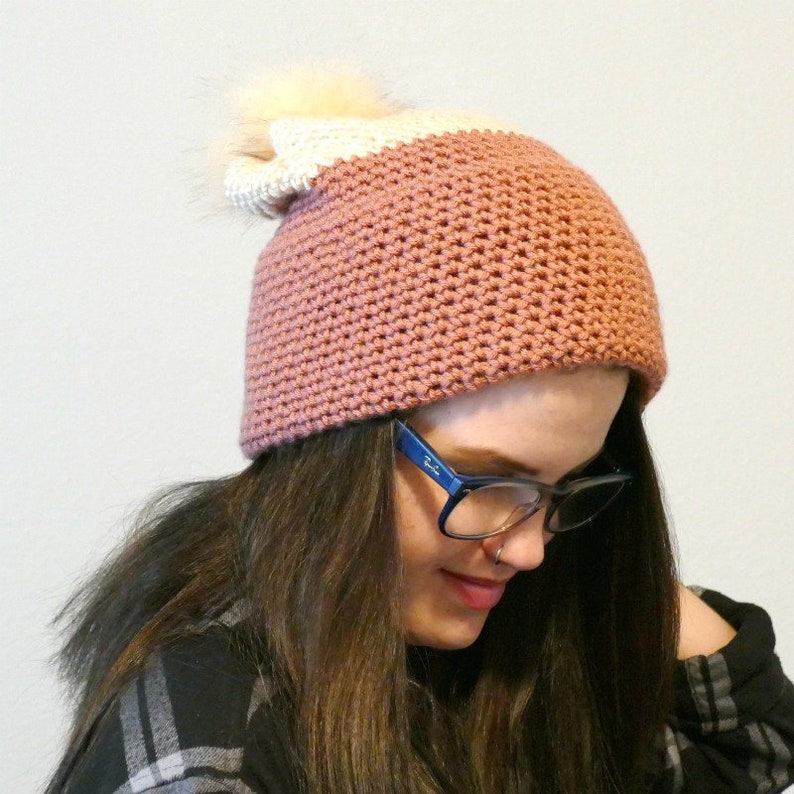 May include: A pink and white crocheted beanie with a fluffy white pom pom. The beanie is worn by a person with long brown hair and glasses.