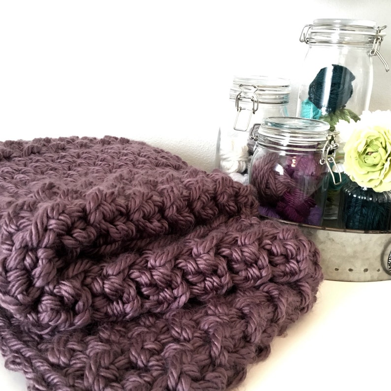 May include: A soft, purple knitted blanket with a textured pattern. The blanket is folded in half and is sitting on a white surface. There are three glass jars with yarn in the background.