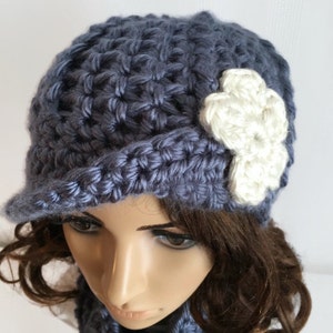 May include: A handmade, blue crochet hat with a small brim and a white flower accent. The hat is made of a thick yarn and has a textured stitch pattern. The hat is shown on a mannequin head.