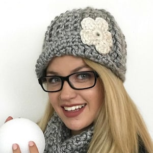 May include: A gray crocheted hat with a cream-colored flower accent. The hat is part of a matching set with a gray scarf. The hat and scarf are made of a thick, textured yarn. The hat is worn by a person wearing black glasses.