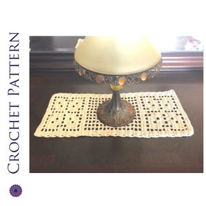 May include: A crocheted beige table runner with a decorative lamp base on top. The lamp base is made of metal with a gold finish and has a clear glass shade.