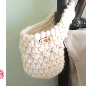 May include: Cream-colored crochet basket with a textured stitch pattern. The basket has a small wooden tag that reads "made with a twist". The words "CROCHET PATTERN" are printed vertically in blue on the left side of the image. The basket is hanging from a dark brown metal object.