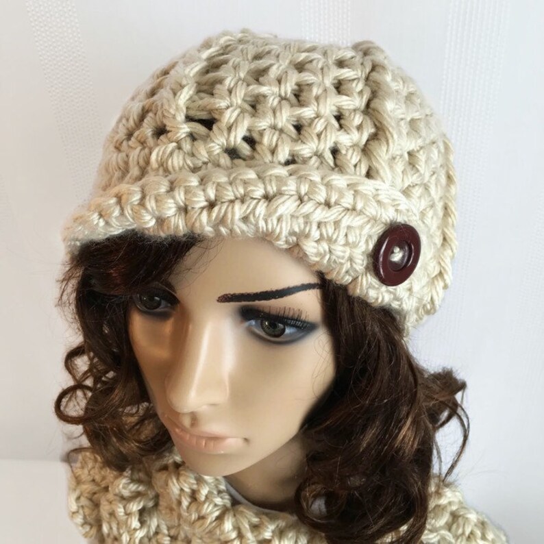 May include: A cream-colored, crocheted hat with a small brim and a large, dark brown button. The hat is part of a set, with a matching scarf visible in the image. The hat is made of thick yarn and has a textured appearance.