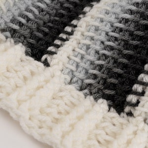 May include: Close-up of a white, gray, and black striped crocheted fabric. The fabric is made with a chunky yarn and has a textured, fuzzy appearance.
