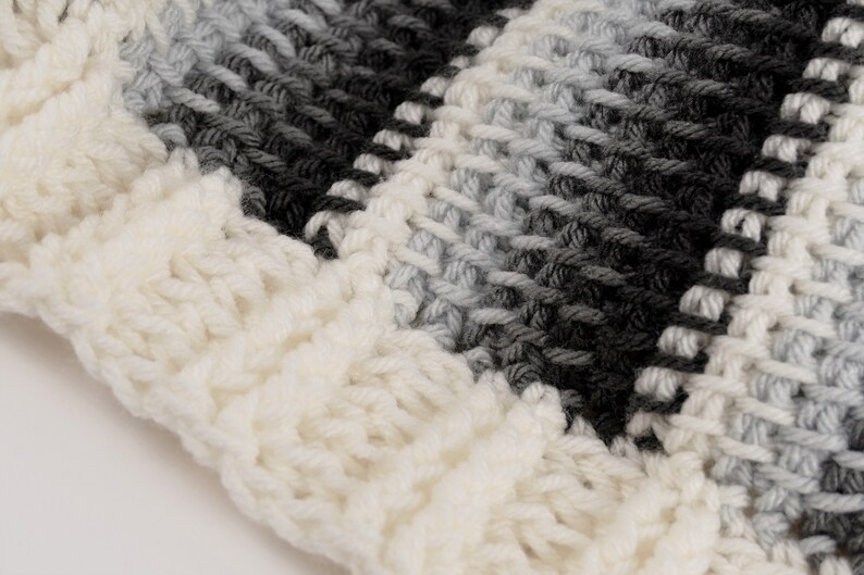 May include: Close-up of a white, gray, and black striped crocheted fabric. The fabric is made with a chunky yarn and has a textured, fuzzy appearance.