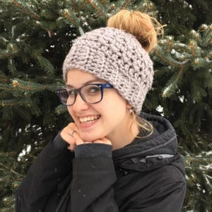 Peut inclure: Une femme portant un bonnet en crochet gris avec un chignon sur le dessus. Le bonnet a un motif textur&eacute; et est tir&eacute; sur ses oreilles. Elle sourit et regarde la cam&eacute;ra.