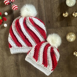 May include: Two handmade crochet beanies with red and white stripes and white faux fur pom-poms. The beanies are in different sizes, suggesting a matching set for adults and children. The hats are on a wooden surface.