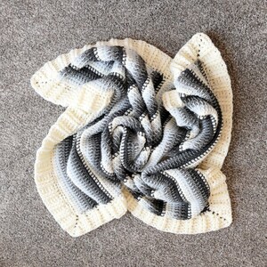 May include: A white and gray striped crocheted blanket with a textured pattern. The blanket is folded and arranged in a circular shape.