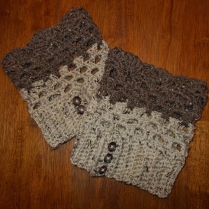 以下が含まれることがあります： 茶色とベージュのクロシェ編みレッグウォーマー2つ。装飾的なステッチパターンが施されています。レッグウォーマーには茶色のボタンが付いています。