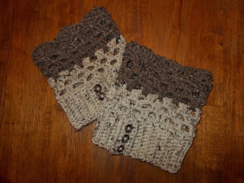 以下が含まれることがあります： 茶色とベージュのクロシェ編みレッグウォーマー2つ。装飾的なステッチパターンが施されています。レッグウォーマーには茶色のボタンが付いています。