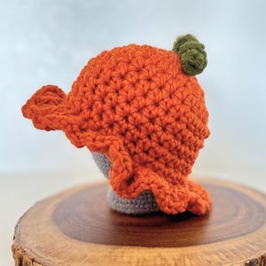 May include: A handmade orange crochet hat, designed to look like a pumpkin, with a green stem. The hat has a ruffled edge and sits on a grey base. The hat is displayed on a wooden surface.