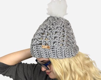 May include: A grey crocheted beanie hat with a white faux fur pom-pom. The hat has a ribbed brim and a wooden tag. The hat is worn by a person with blonde hair and blue glasses.