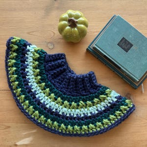 May include: A crocheted neck warmer in navy blue, green, and light blue. The warmer is made with a variety of stitches and has a scalloped edge.
