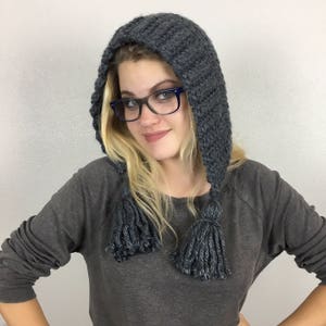 Könnte beinhalten: Eine graue Kapuze mit langen Quasten. Die Mütze ist gestrickt und hat eine lockere Passform. Das Model trägt die Mütze und hat blonde Haare und trägt eine Brille.