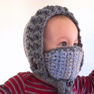 May include: A gray and blue crocheted baby hood with a face covering. The hood has a textured pattern and a button closure. The face covering is blue and gray. The baby is wearing a red and white striped shirt.