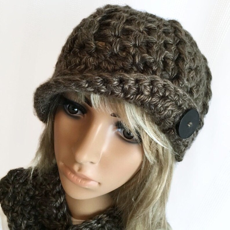 May include: A brown crocheted hat with a small brim and a black button detail. The hat is made of a textured yarn and appears to be handmade. The hat is part of a set, with a matching scarf visible in the image.