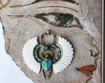 May include: A gold scarab beetle pendant with turquoise and blue accents, set against a white background with wings. The pendant is a detailed representation of an Egyptian scarab beetle.