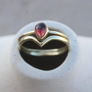 Könnte beinhalten: Ein goldener Ringsatz mit einem tropfenförmigen, roten Edelstein. Der Ring liegt auf einer weißen, strukturierten Oberfläche. Das Band hat ein Chevron-Design. Der Ring ist ein Schmuckstück.
