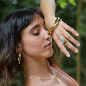 Puede incluir: Una mujer lleva un anillo de oro con una piedra ovalada azul claro, tres pulseras de oro y pendientes de aro de oro. Lleva un top de tirantes color óxido.