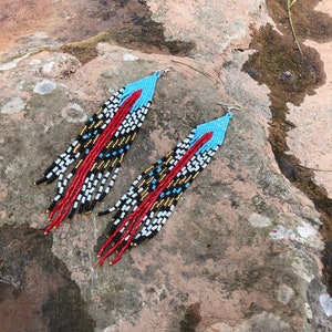 Puede incluir: Un par de aretes largos y abalorios con un patrón geométrico en rojo, blanco, negro, dorado y azul. Los aretes cuelgan de una superficie rocosa.