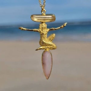 Puede incluir: Un collar de oro con un colgante que representa una figura de sirena sosteniendo una piedra preciosa rosa. El colgante está suspendido de una cadena de oro.
