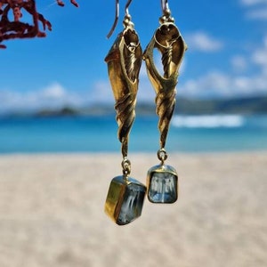 Puede incluir: Pendientes de oro con diseño de concha marina y gotas de piedras preciosas azules. Los pendientes cuelgan de una rama contra un fondo de cielo azul y playa.