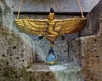 May include: A gold-toned necklace featuring a winged figure pendant with outstretched wings, accented by small dark stones at the top and sides, and a large, faceted, translucent blue teardrop-shaped stone hanging from the bottom center.