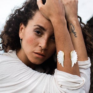 以下が含まれることがあります： 茶色の巻き毛の女性が白いシャツを着て、手首に白い天使の羽根をつけています。彼女は頭を手に持って座り、うつむいています。